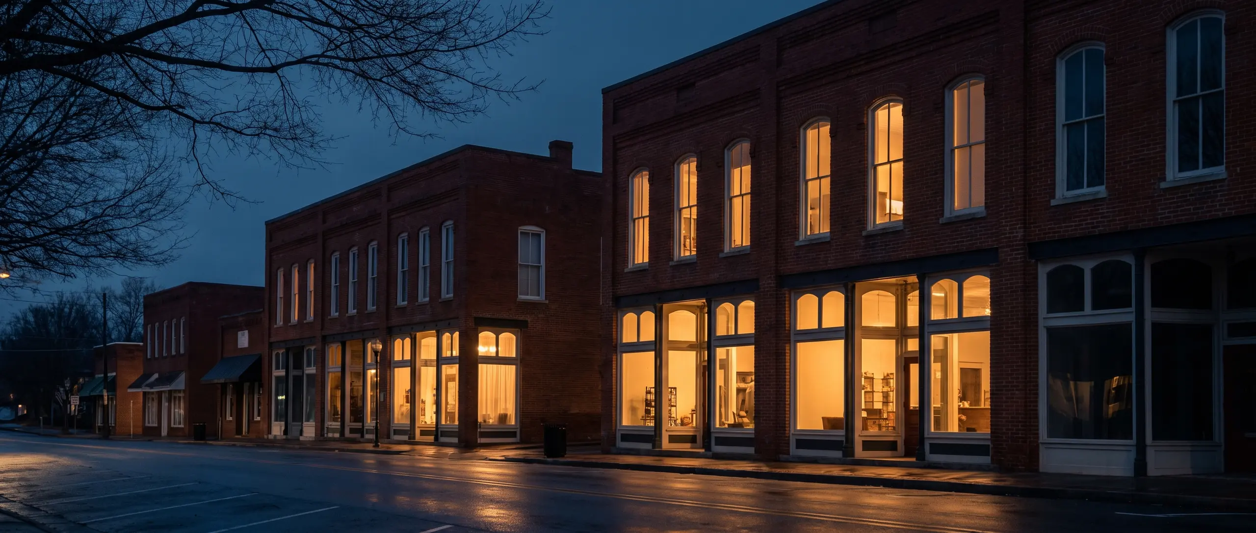 A small-town main street of two-storey brick storefronts at dusk, their ground-floor windows lit warm against a deep blue sky.